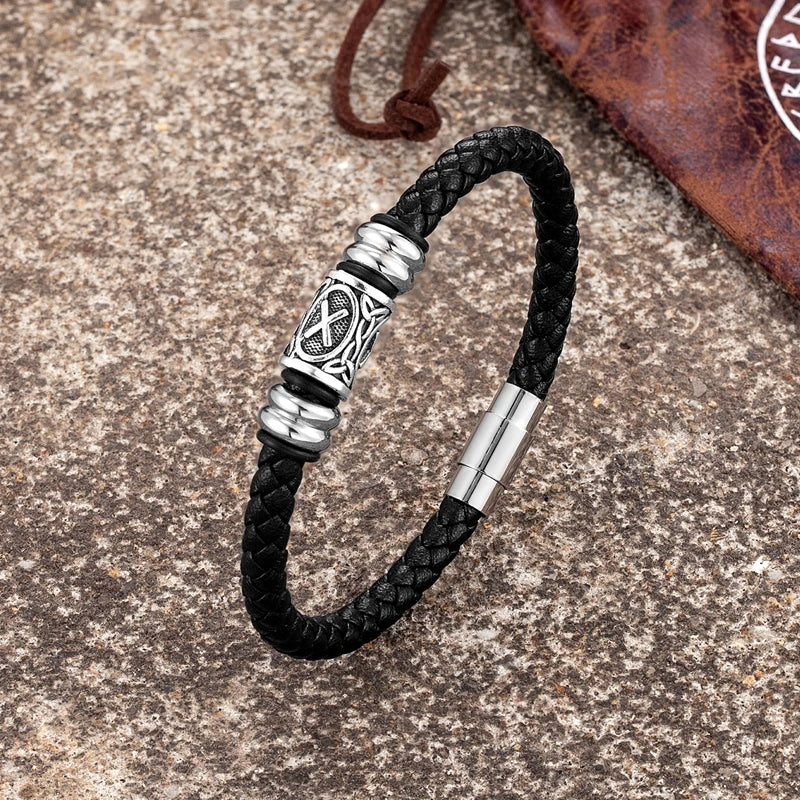 Image of a stainless steel and leather bracelet with a bead featuring an engraved Elder Futhark rune, set against a natural outdoor background.